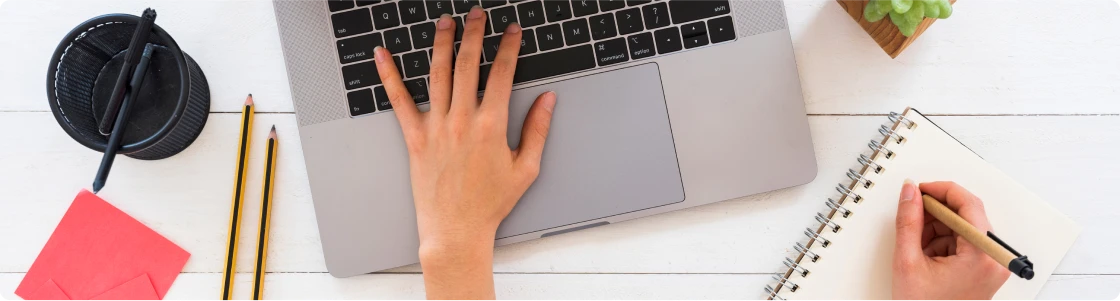 Vista aérea de un escritorio blanco con una laptop abierta, una mano tecleando, y otra mano escribiendo en un cuaderno de notas, acompañado de lápices y decoración de oficina